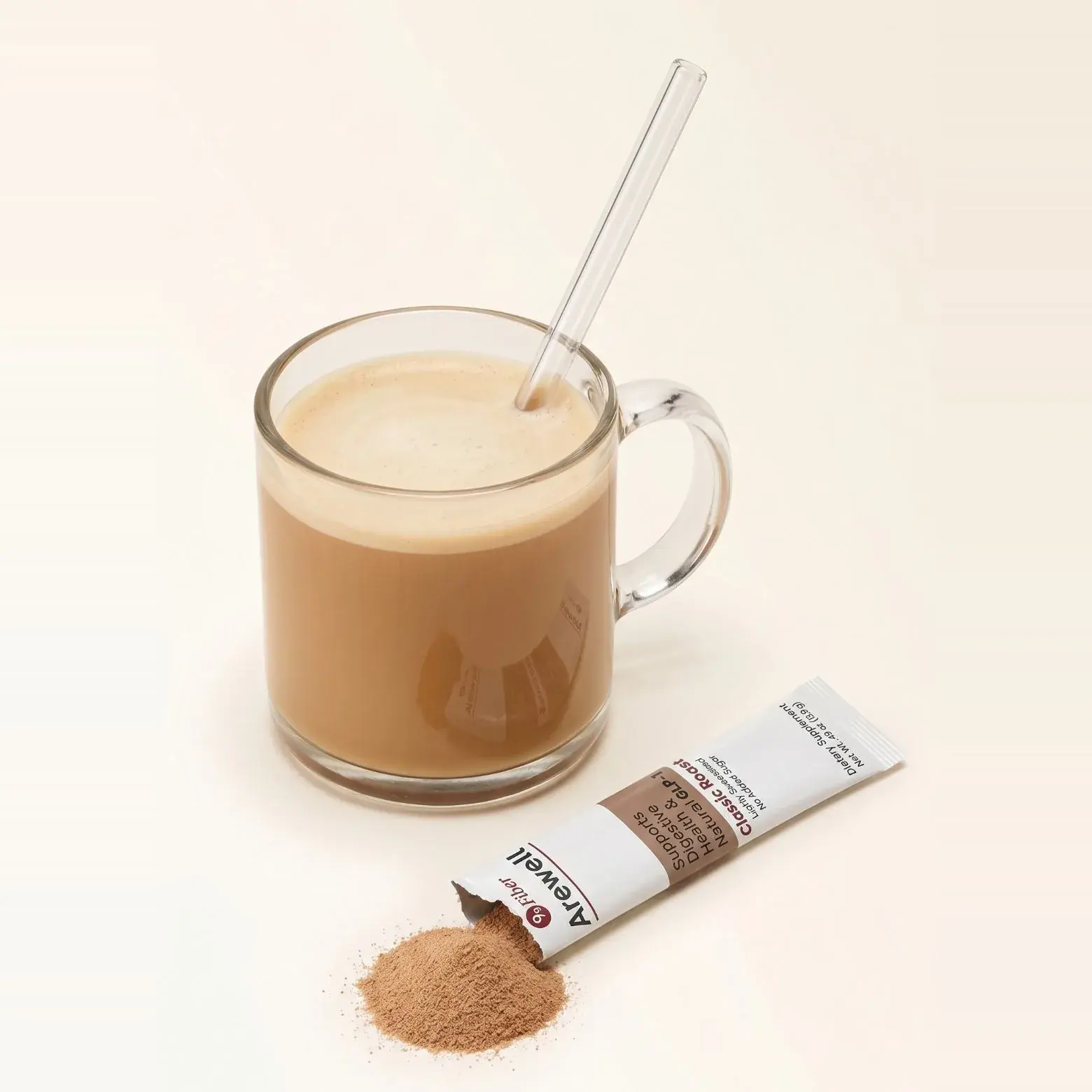 A glass mug of Arewell coffee made with milk, with a glass straw, on a plain cream surface. An opened Classic Roast stick pack lies beside it with the coffee powder spilling out.